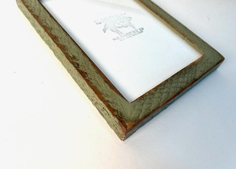 May include: A rectangular picture frame with a weathered, sage green finish and visible wood grain texture. The frame contains a white insert featuring the text SIGNED NUMBERED in black ink, accompanied by a small graphic of a pig illustration.