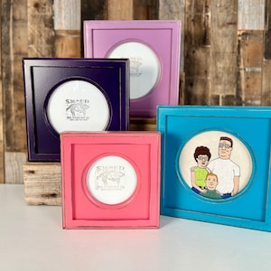 May include: Four different colored picture frames with various images inside. The frames are all square and have a distressed wood finish. The frames are arranged in a row from left to right. The first frame is purple, the second is pink, the third is blue, and the fourth is light blue. The images inside the frames are a black and white drawing of a dog, a black and white drawing of a dog, a cartoon image of a family, and a cartoon image of a family.