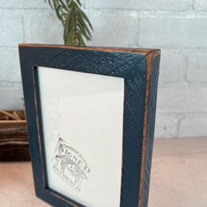K&ouml;nnte beinhalten: Ein dunkelblauer Bilderrahmen mit einer abgenutzten Holzoptik. Der Rahmen hat einen wei&szlig;en Passepartout und ein kleines, graues, gepr&auml;gtes Bild im Inneren. Das Bild sagt "SIGNED" mit einer kleinen Illustration eines Hundes.
