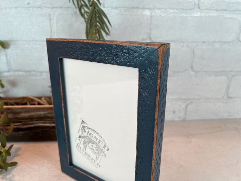 K&ouml;nnte beinhalten: Ein dunkelblauer Bilderrahmen mit einer abgenutzten Holzoptik. Der Rahmen hat einen wei&szlig;en Passepartout und ein kleines, graues, gepr&auml;gtes Bild im Inneren. Das Bild sagt "SIGNED" mit einer kleinen Illustration eines Hundes.
