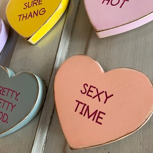May include: Five wooden heart-shaped signs with different sayings. The signs are painted in various colors, including yellow, purple, pink, peach, and gray. The sayings on the signs are "Sure Thang", "Hot", "Pretty, Pretty Good", "Sexy Time", and "Ever".