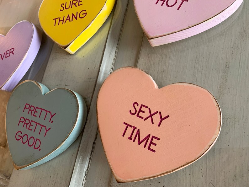 May include: Five wooden heart-shaped signs with different sayings. The signs are painted in various colors, including yellow, purple, pink, peach, and gray. The sayings on the signs are "Sure Thang", "Hot", "Pretty, Pretty Good", "Sexy Time", and "Ever".