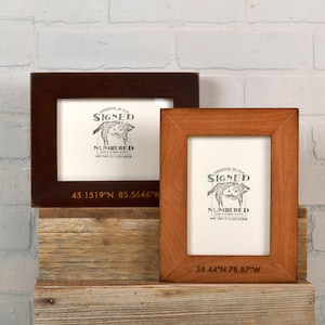 May include: Two wooden picture frames with a white background and black text. The top frame is brown and the bottom frame is light brown. Both frames have the text "Signed and Numbered" with a small image of a pig. The top frame has the coordinates "45.1519'N 85.5646'W" and the bottom frame has the coordinates "38.44'N 78.87'W".