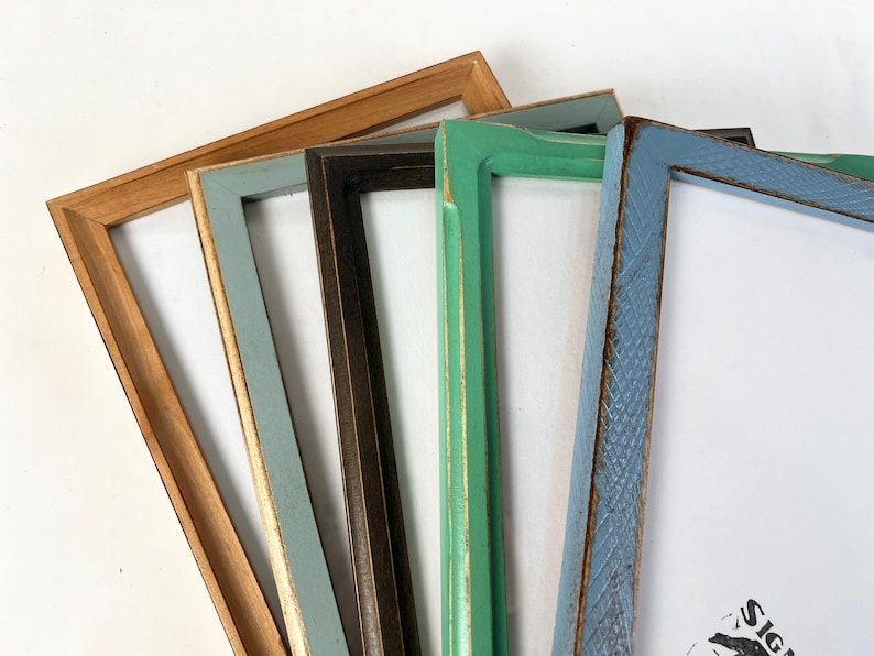 May include: A collection of five rectangular picture frames arranged in a fan shape. The frames feature various finishes including natural wood, matte light blue, dark brown, mint green, and a textured light blue with visible wood grain patterns.