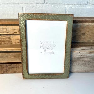 May include: A rectangular picture frame with a distressed sage green finish and visible wood grain texture. The frame surrounds a white insert featuring a centered graphic of a pig with the text "SIGNED" above and "NUMBERED" below.