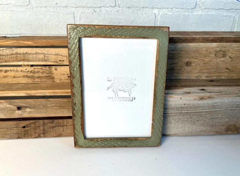May include: A rectangular picture frame with a distressed sage green finish and visible wood grain texture. The frame surrounds a white insert featuring a centered graphic of a pig with the text "SIGNED" above and "NUMBERED" below.