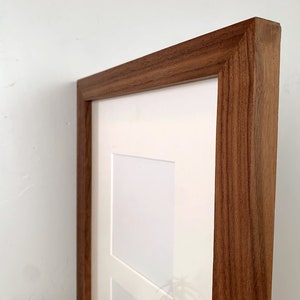 May include: Close-up of a wooden picture frame with a natural wood grain finish. The frame has a white matting and is designed to hold multiple photos or artwork. The frame is displayed against a white wall.