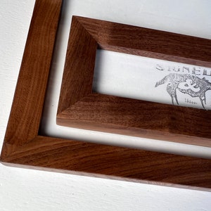 May include: Two wooden picture frames with a rich brown finish are stacked on a white surface. The frames are rectangular, with a visible print inside the smaller frame. The print features a wolf illustration and the words "SIGNED &" in a vintage style.
