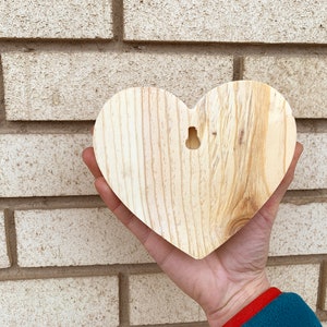 May include: A plain wooden heart-shaped ornament with a hole for hanging. The heart is light brown and has a natural wood grain.