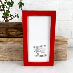May include: A rectangular red picture frame with a white interior and a printed image of a wolf. The frame is placed in front of a wooden box and a small potted plant. The text "SIGNED NUMBERED" is printed on the image.