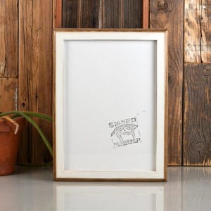 May include: A rectangular wooden picture frame with a white mat. The frame has a light brown finish and a white interior. The image has a stamp that says "SIGNED NUMBERED". A small terracotta pot with a green plant is to the left of the frame.