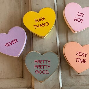 May include: Five wooden heart-shaped decorations with different sayings. The hearts are painted in various colors, including yellow, pink, purple, gray, and peach. The sayings on the hearts are "Sure Thang", "Ur Hot", "4ever", "Pretty, Pretty Good", and "Sexy Time".