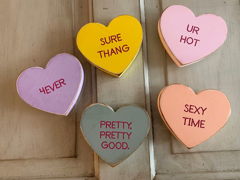 May include: Five wooden heart-shaped decorations with different sayings. The hearts are painted in various colors, including yellow, pink, purple, gray, and peach. The sayings on the hearts are "Sure Thang", "Ur Hot", "4ever", "Pretty, Pretty Good", and "Sexy Time".