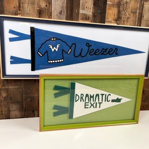 May include: A blue felt pennant featuring a graphic of a sweater with a W logo and the text Weezer, framed in a dark blue frame. Below it, a white felt pennant with the text DRAMATIC EXIT and a pointing hand, framed in a green frame.