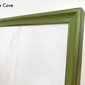 May include: A rectangular picture frame with a matte olive green finish and visible distressed edges. The frame features a beveled inner edge and contains a white mat board with a faint botanical print. The text Foxy Cove is printed in black.
