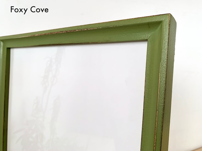 May include: A rectangular picture frame with a matte olive green finish and visible distressed edges. The frame features a beveled inner edge and contains a white mat board with a faint botanical print. The text Foxy Cove is printed in black.
