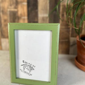 May include: A rectangular picture frame with a distressed light green finish. The frame features a white insert with a black graphic logo in the bottom left corner that depicts a pig and the text Signed Numbered.