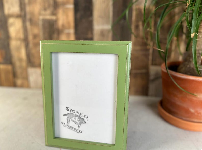 May include: A rectangular picture frame with a distressed light green finish. The frame features a white insert with a black graphic logo in the bottom left corner that depicts a pig and the text Signed Numbered.