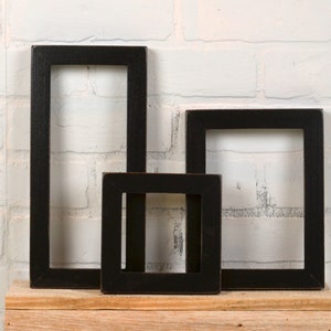 May include: Three black picture frames of different sizes are arranged on a wooden shelf against a white brick wall. The frames are all empty and have a simple, rustic design.