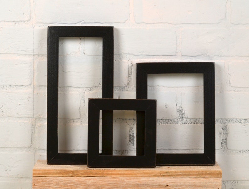 May include: Three black picture frames of different sizes are arranged on a wooden shelf against a white brick wall. The frames are all empty and have a simple, rustic design.
