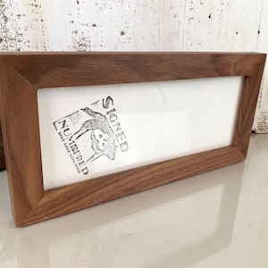 May include: A rectangular wooden picture frame with a dark brown finish. The frame holds a white insert with a black and white graphic and the words "SIGNED NUMBERED SALT LAKE CITY, UTAH". The frame is angled slightly.