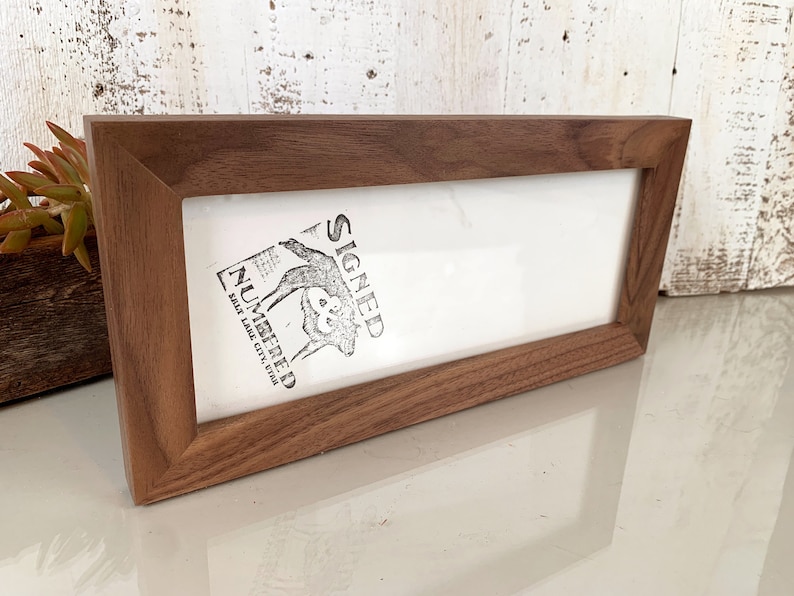 May include: A rectangular wooden picture frame with a dark brown finish. The frame holds a white insert with a black and white graphic and the words "SIGNED NUMBERED SALT LAKE CITY, UTAH". The frame is angled slightly.