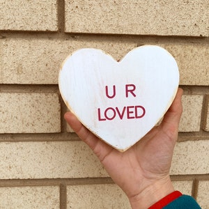 May include: White wooden heart-shaped sign with the words "U R LOVED" in red lettering.