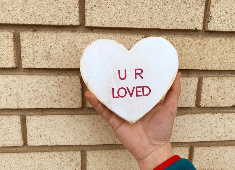 May include: White wooden heart-shaped sign with the words "U R LOVED" in red lettering.