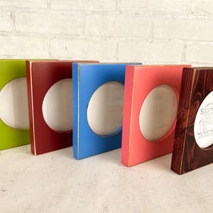 May include: A collection of five colorful wooden picture frames. The frames are painted in green, red, blue, pink, and dark brown. Each frame has an oval opening for a photo. The dark brown frame has the word "SIGNED" visible.