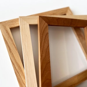 May include: Three wooden picture frames of different sizes and wood tones. The frames are stacked on top of each other, with the largest frame on the bottom and the smallest frame on top. The frames are all made of natural wood and have a simple, elegant design.