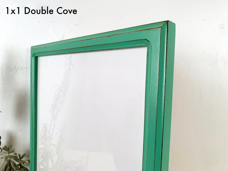 May include: A rectangular picture frame with a mint green finish and distressed dark brown edges. The frame features a double cove profile and includes the text 1x1 Double Cove in the upper left corner, surrounding a blank white center.