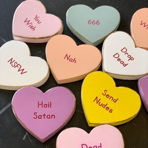 May include: A collection of wooden heart-shaped candy conversation hearts with various messages, including "You Wish", "666", "Nah", "Drop Dead", "NSFW", "Hail Satan", "Send Nudes", and "Dead Inside".