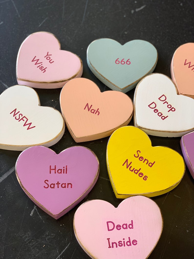 May include: A collection of wooden heart-shaped candy conversation hearts with various messages, including "You Wish", "666", "Nah", "Drop Dead", "NSFW", "Hail Satan", "Send Nudes", and "Dead Inside".