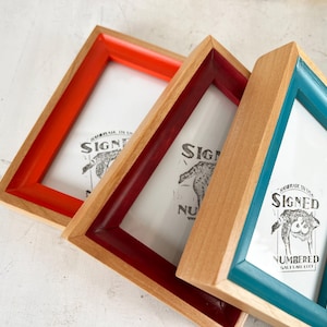 May include: Three wooden picture frames with colorful accents. One frame has a red border, another a burgundy border, and the third a teal border. Each frame contains a white print with the words "SIGNED" and "NUMBERED".
