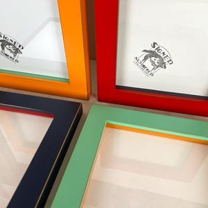 May include: A collection of colorful picture frames in orange, red, navy blue, and mint green. Each frame has a white interior and a logo in the upper left corner. The frames are arranged at various angles, showcasing their vibrant colors.