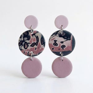 Op de afbeelding: Een paar oorhangers met een ontwerp van drie cirkels. De bovenste en onderste cirkels zijn effen mauve van kleur, terwijl de middelste cirkel een zwart, wit en mauve gemarmerd patroon heeft. De oorbellen zijn verbonden met zilverkleurige ringetjes.