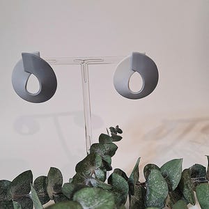 May include: A pair of gray, abstract-shaped earrings with a teardrop cutout, displayed on a clear stand. The earrings have a modern design and are set against a white background. Green eucalyptus leaves are in the foreground.