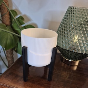May include: White ceramic planter with a ribbed texture, sitting on a black metal stand. The stand has three legs and is on a wooden surface.