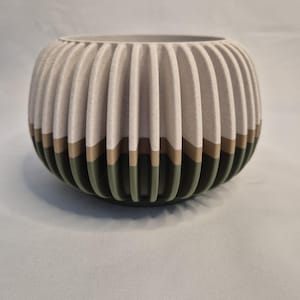 May include: A round, ribbed planter in neutral tones. The upper part is light beige, with a brown band and an olive green base. The vertical ribs give a textured appearance. The planter is suitable for indoor use.
