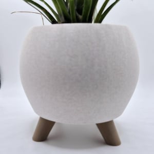 May include: A white, round planter with three brown wooden legs, holding a green plant with long, slender leaves. The planter has a textured surface and a modern design, suitable for indoor decor.