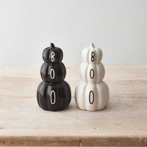 May include: Two stacked pumpkin figurines, one black and one white, each featuring the word BOO written vertically in contrasting lettering across the three rounded pumpkin segments. The ceramic objects have a smooth, glossy finish and rounded shapes.