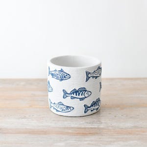 May include: White ceramic cylindrical planter decorated with blue fish illustrations. The planter has a smooth, unglazed finish and is suitable for small plants or as a decorative item. The fish designs vary in style.