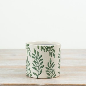 May include: A cylindrical ceramic planter featuring a cream-coloured base with a crackle glaze finish. The exterior is decorated with a repeating pattern of green leafy branches that extend vertically around the entire circumference of the vessel.