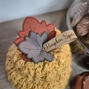 May include: A small, round, yellow pumpkin-shaped decoration with a textured surface. It features a decorative leaf and a tag that reads "Pumpkin Spice". The leaf is made of brown and grey felt. The decoration is likely for autumn decor.