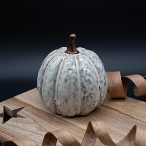 May include: A ceramic pumpkin sculpture with a glossy, mottled off-white and light brown glaze, featuring deep vertical ridges and a smooth, dark brown stem. The pumpkin has a rounded, segmented shape and a textured surface finish.