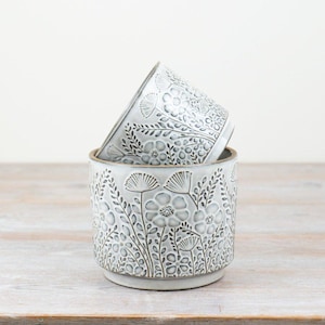 May include: Two cylindrical ceramic planters stacked vertically, featuring a light grey glaze with a dark grey embossed floral pattern of wildflowers and stems. The planters have a smooth rim and a slightly recessed base, showcasing a detailed botanical relief texture across the exterior surface.