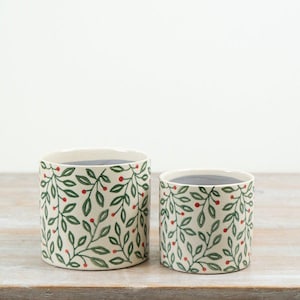 May include: Two cylindrical ceramic planters with a cream-coloured base featuring a repeating hand-painted pattern of green leafy vines and small red berries. The planters have a smooth finish and a dark grey interior, displayed side by side.
