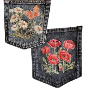 May include: Two dark denim pockets featuring embroidered designs. One pocket displays white daisies and an orange butterfly. The other pocket features a cluster of red poppies with green stems and leaves. Both pockets have visible stitching.