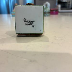 May include: A small, square, white trinket box with green trim and a gold-coloured top. The box has the text "Peint Main Limoges France NG" in black script. The box is sitting on a light-coloured surface.