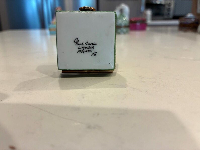 May include: A small, square, white trinket box with green trim and a gold-coloured top. The box has the text "Peint Main Limoges France NG" in black script. The box is sitting on a light-coloured surface.
